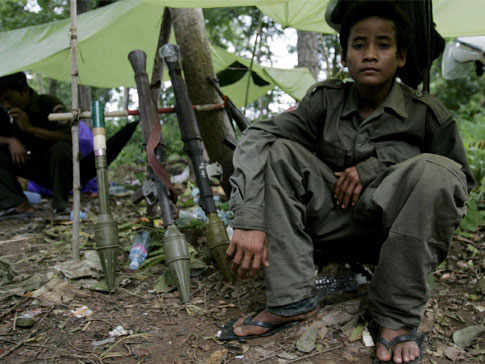 niño sentado en el suelo en un campamento, a su lado armas